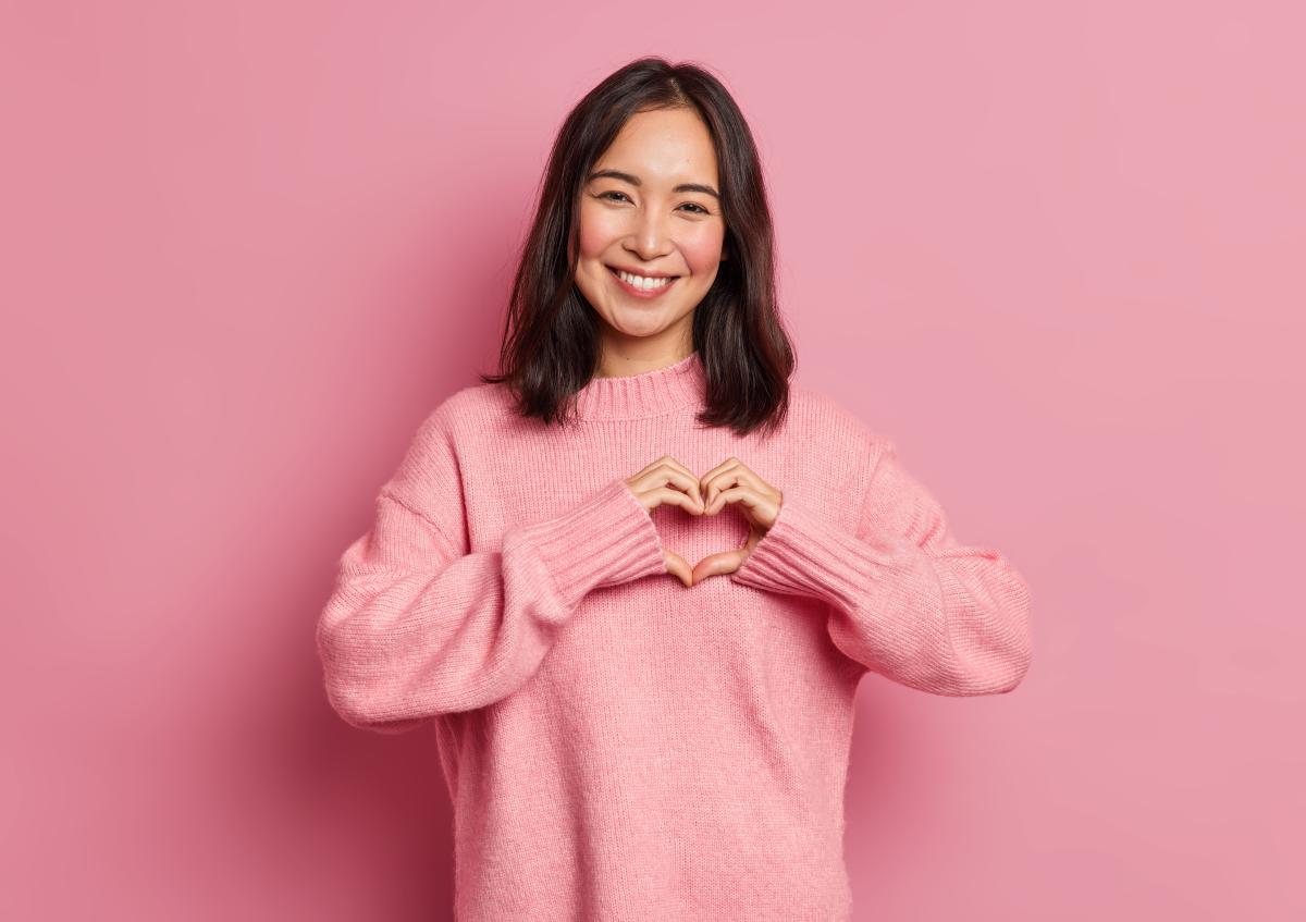 A smiling woman looking forward, making a heart shape with her hands in front of her chest.