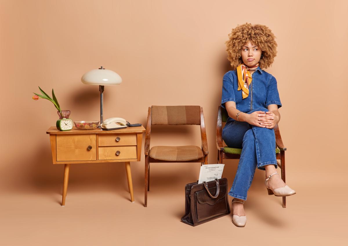 A young woman sitting on a chair next to an empty chair and a small table with a lamp, telephone, clock, and flower vase, looking away with an expression of frustration.