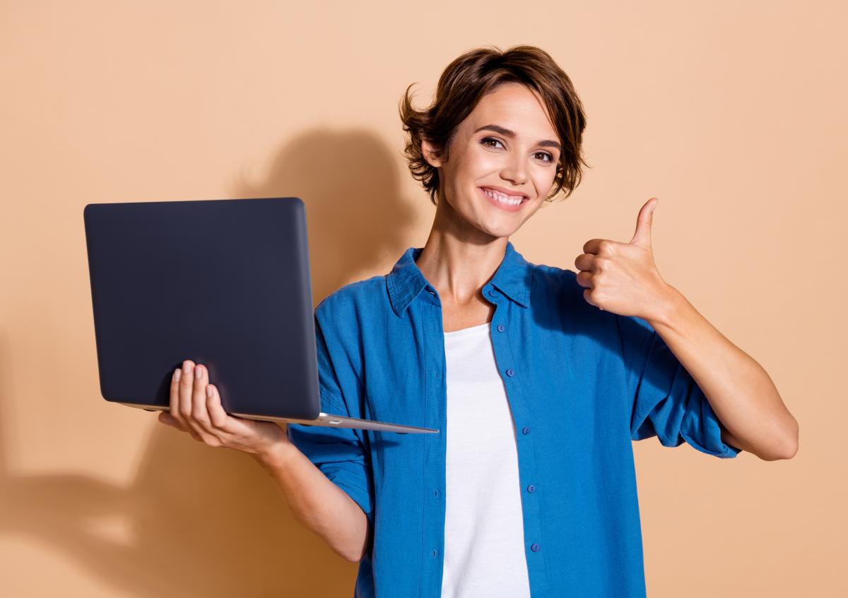 Vrouw lacht vrolijk en kijkt tevreden in de camera. Ze houdt een laptop in haar linkerhand en steekt met haar rechterhand een duim omhoog.
