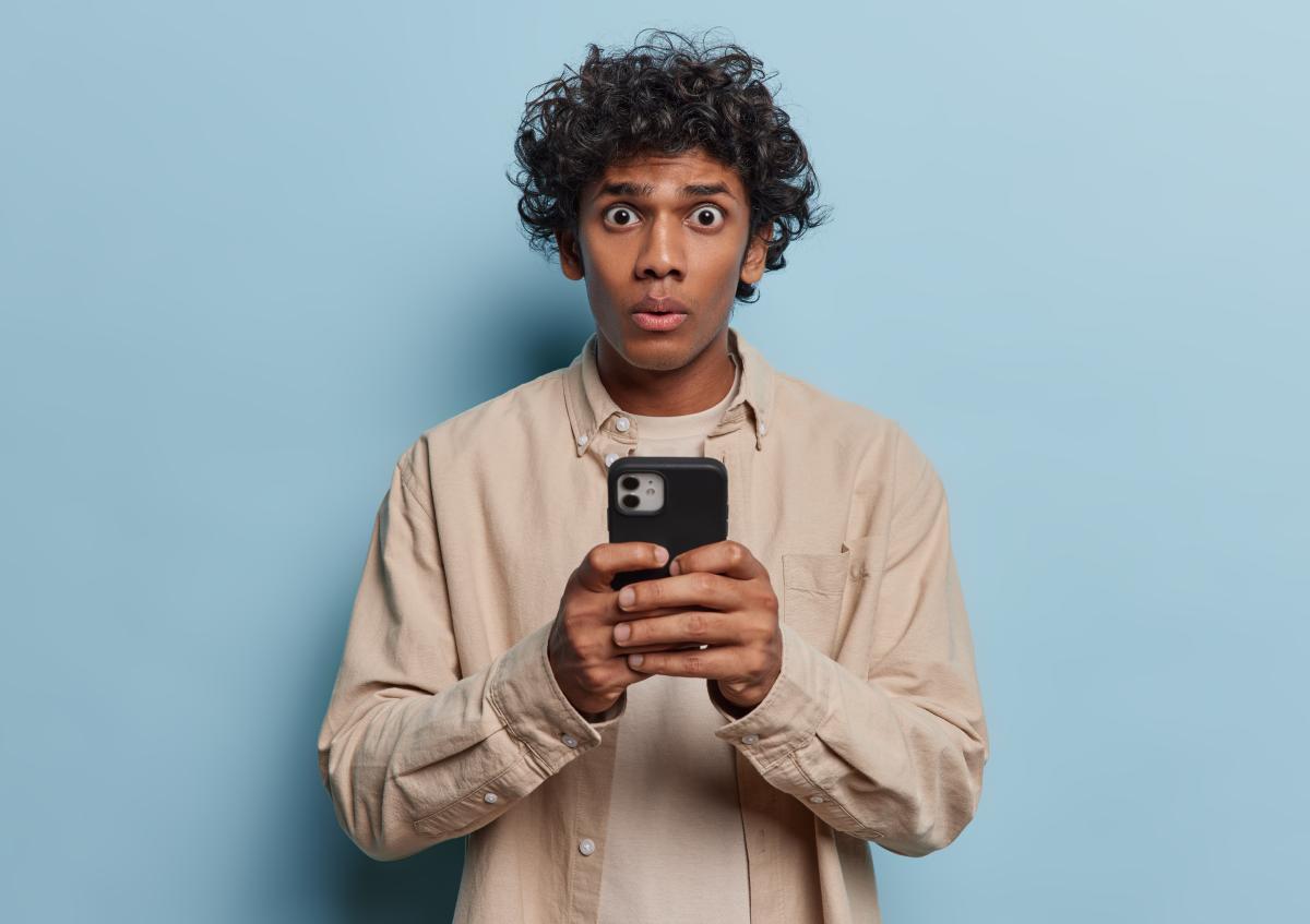 Een jonge man houdt met beide handen een telefoon vast. Hij kijkt er met grote ogen en samengeknepen lippen naar, en ziet er verrast of geschrokken uit.