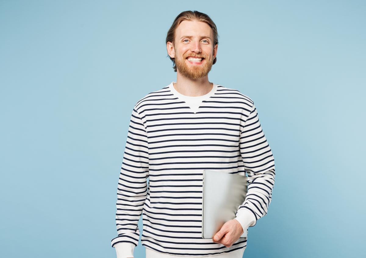 Man kijkt vriendelijk en ontspannen naar de camera met een brede glimlach. Hij houdt een laptop met één hand dicht tegen zijn lichaam.