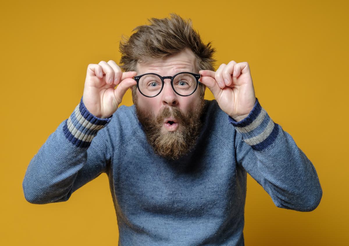 A surprised man with a full beard and tousled hair looks directly at the camera. He is holding black-rimmed round glasses up to his face with both hands