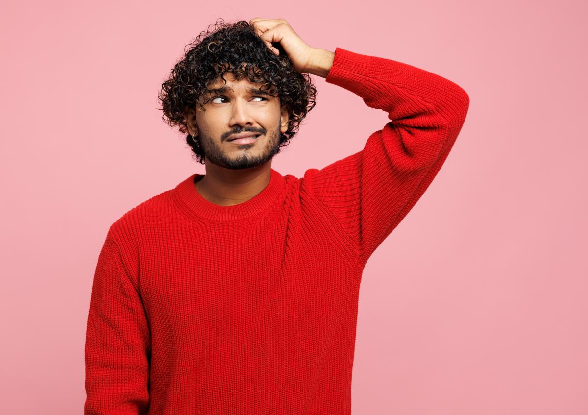 Een verwarde jonge man krabt met zijn linkerhand aan zijn hoofd en kijkt iets naar rechts met een vragende blik.
