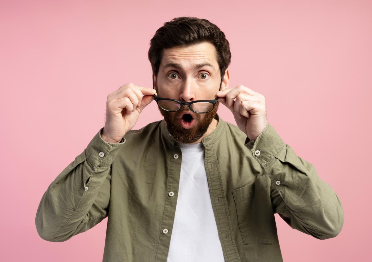 A man with a beard is holding his glasses down from his eyes, looking surprised and shocked, with his mouth open as if saying "wow" or expressing amazement.