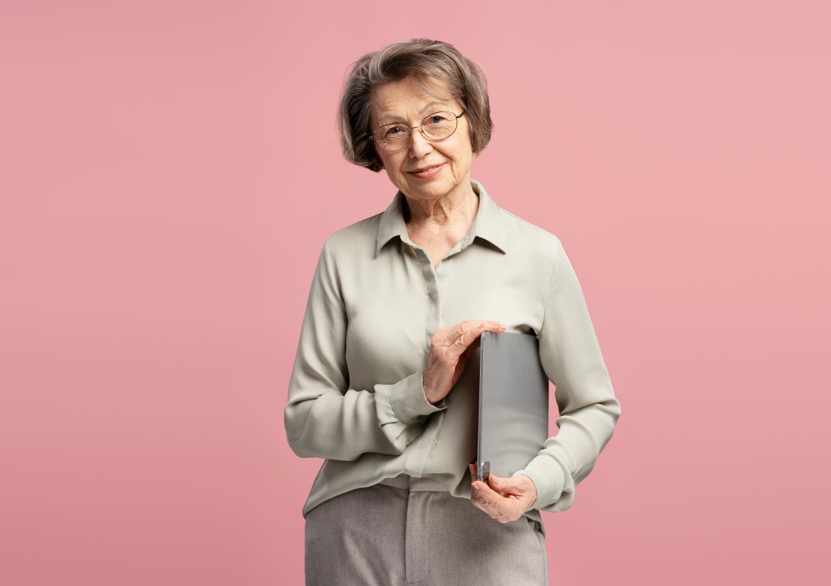 An elderly woman gently smiling, holding a closed laptop close to her chest with both hands.