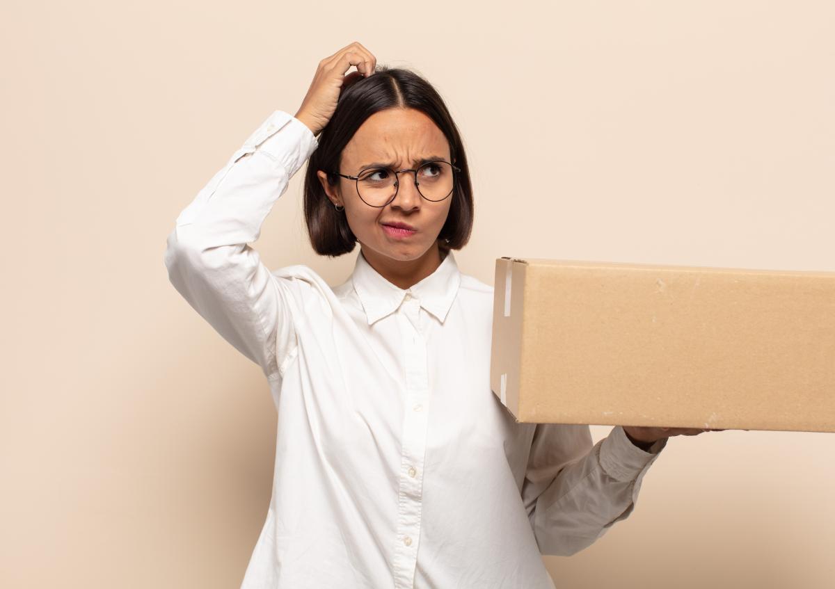 A woman with a frown and a puzzled look scratches her head while holding a large cardboard box in one hand, as if she doesn’t understand something or is deep in thought