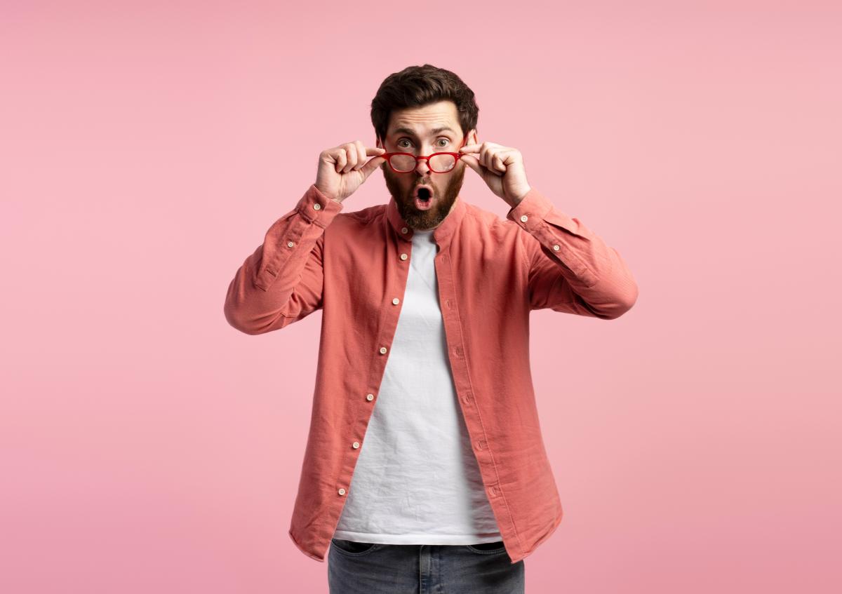 A man with a beard is holding his glasses down from his eyes, looking surprised and shocked, with his mouth open as if saying "wow" or expressing amazement.