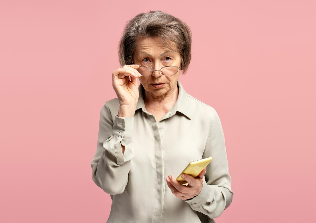 An elderly woman holding a smartphone in one hand while adjusting her glasses with the other, looking directly at the camera with a serious and focused expression.