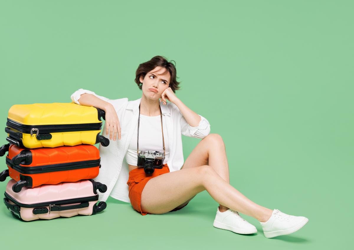 Vrouw zit op de grond met haar hand tegen haar wang, terwijl ze naast een stapel koffers zit. Ze kijkt omhoog met een frons en een lichte zucht, wat een uitdrukking van verveling of ongeduld toont.