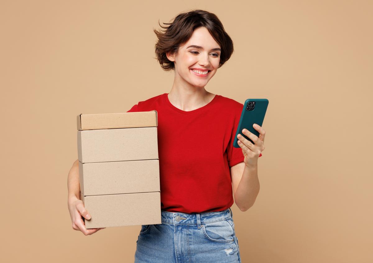 A young woman smiles while holding a stack of four cardboard boxes in one arm and looking at her smartphone in the other hand