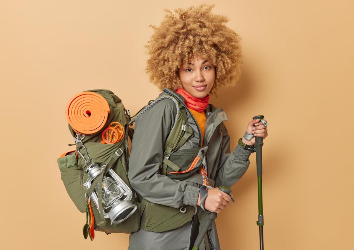 A confident young woman with camping gear, including a large backpack and trekking poles, looking at the camera with a slight smile.