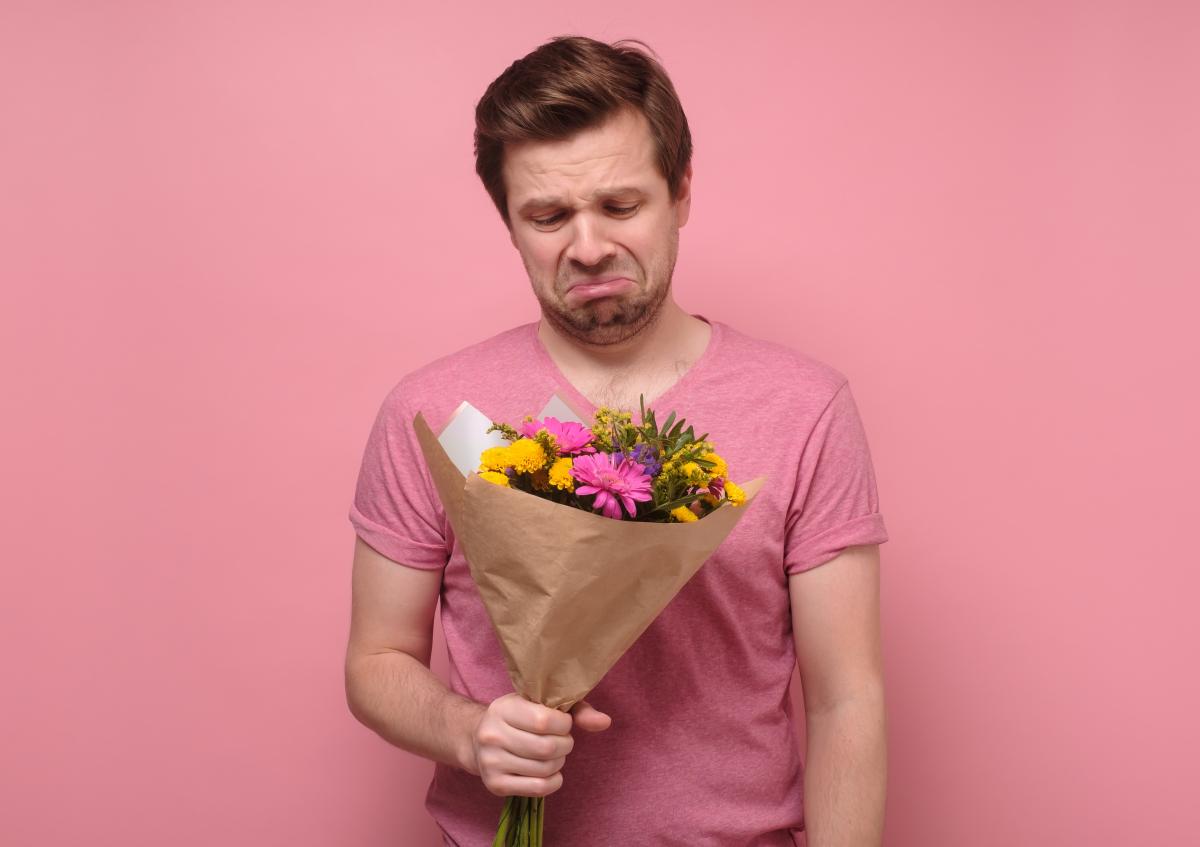 A man holding a bouquet of flowers wrapped in paper. He looks at the flowers with a sad, disappointed expression, his lips turned downwards and eyebrows furrowed