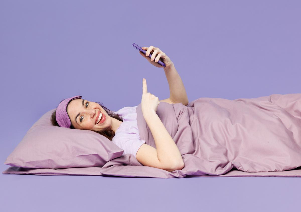 A woman lying in bed, smiling, pointing upward with one hand while holding a phone with the other. She is wearing a sleep mask pushed up on her forehead.