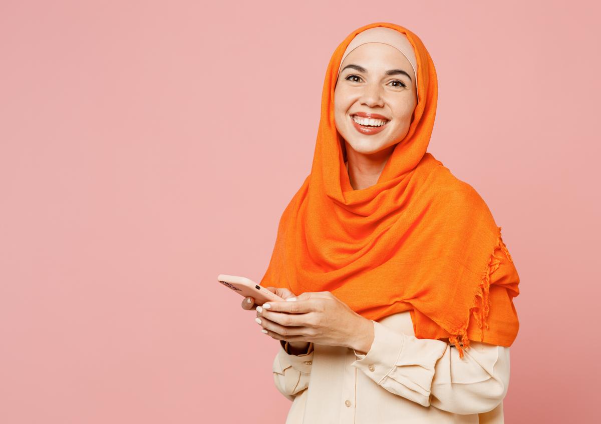 A smiling woman wearing a headscarf looks down at her phone while holding it with both hands, appearing happy and engaged.