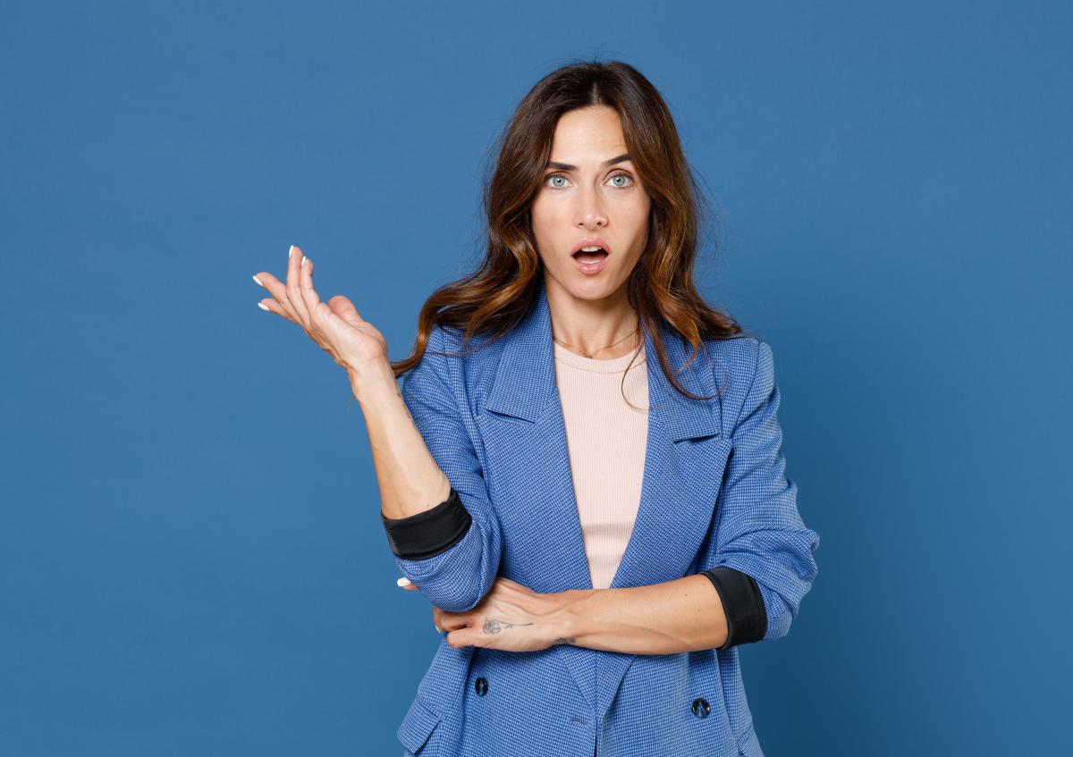 Vrouw kijkt verbaasd met open mond en opgetrokken wenkbrauwen, terwijl ze haar rechterhand opwerpt en haar linkerarm gekruist houdt.