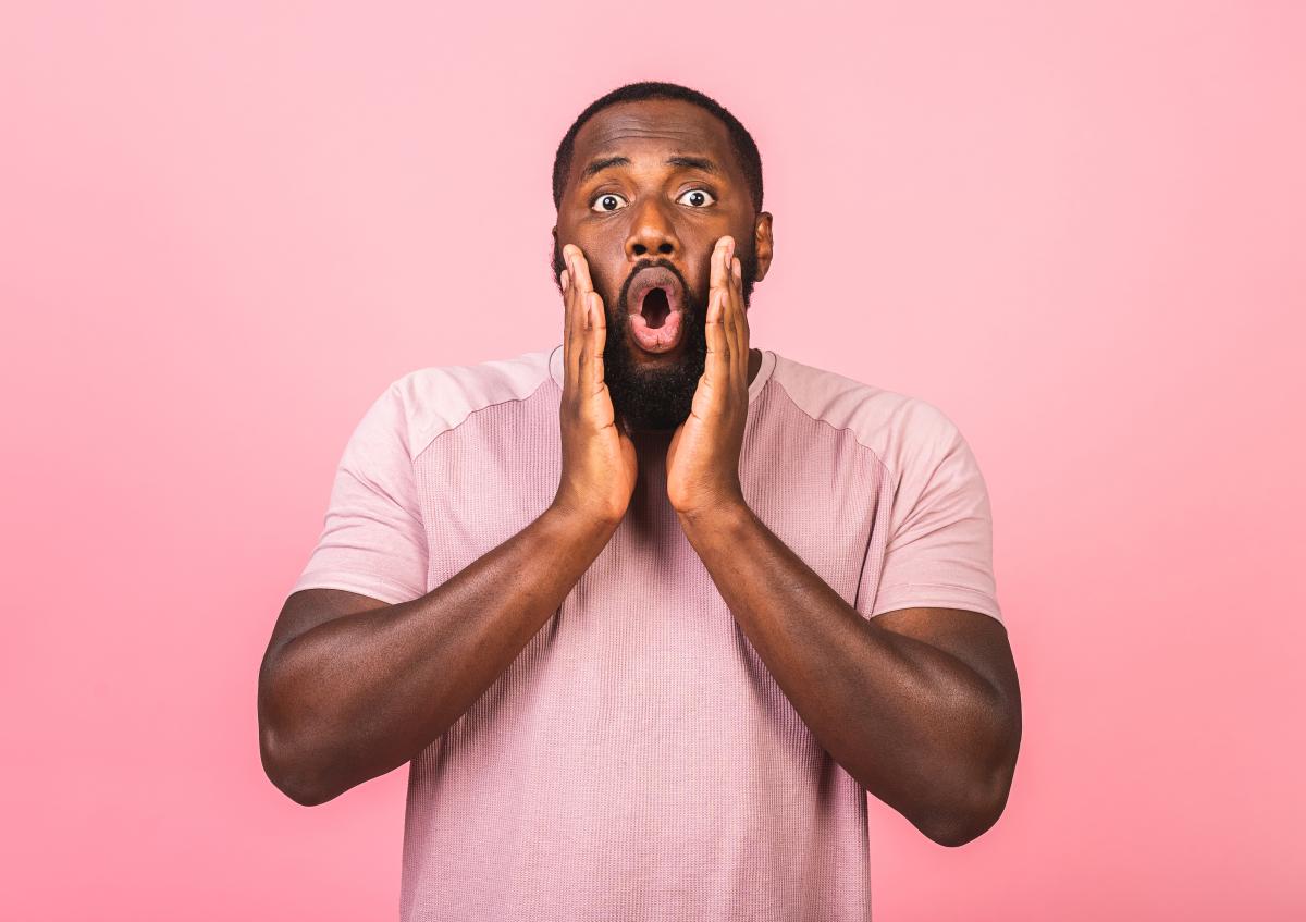 A young man expresses surprise and shock, with his mouth open and hands raised near his face.