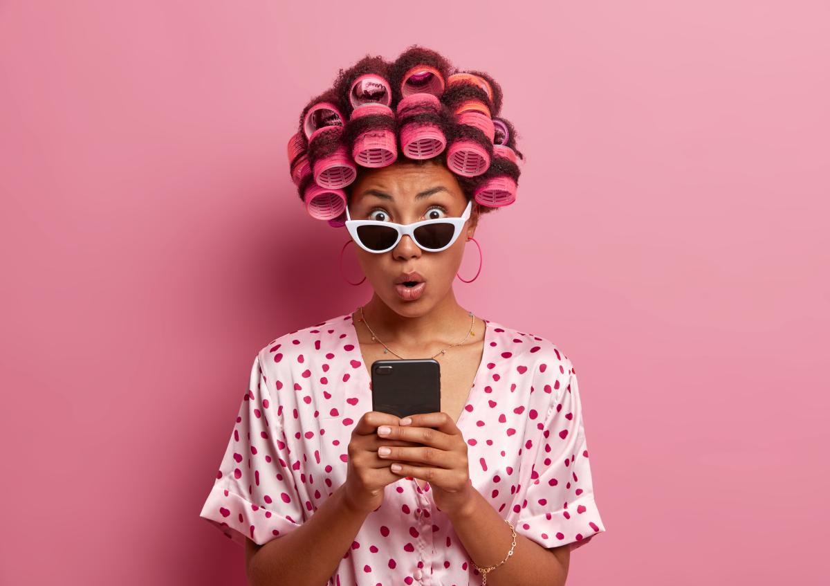 Vrouw kijkt met grote ogen en open mond verbaasd naar haar telefoon. Ze draagt een bril en heeft haar haar in krulspelden.