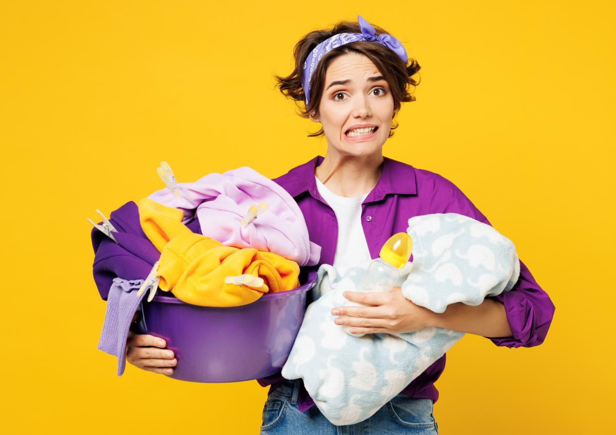 A young woman holds a laundry basket filled with clothes. Her facial expression shows anxiety or stress as she bites her lip.