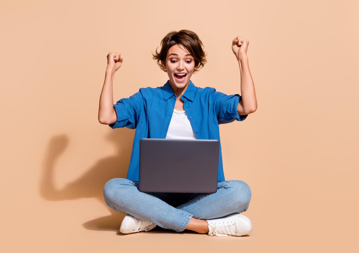 Vrouw in kleermakerszit met laptop op schoot, verheugd en juigend terwijl ze naar het scherm kijkt