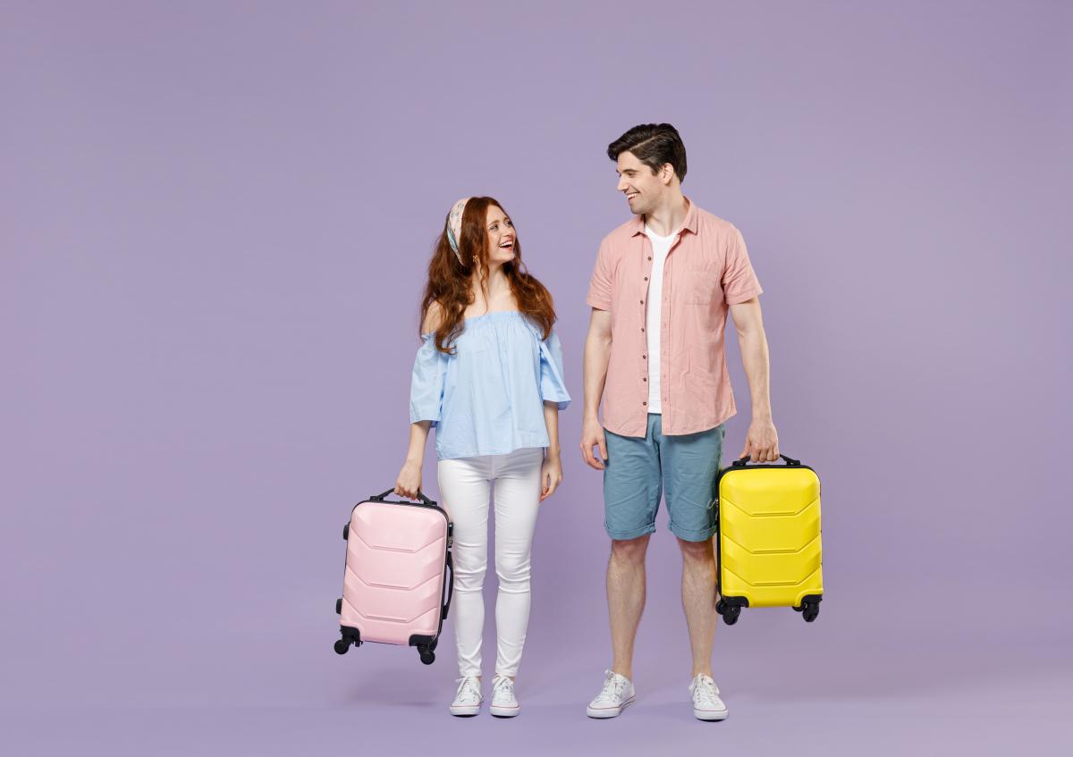 A young man and woman, each holding a small suitcase while smiling and looking at each other
