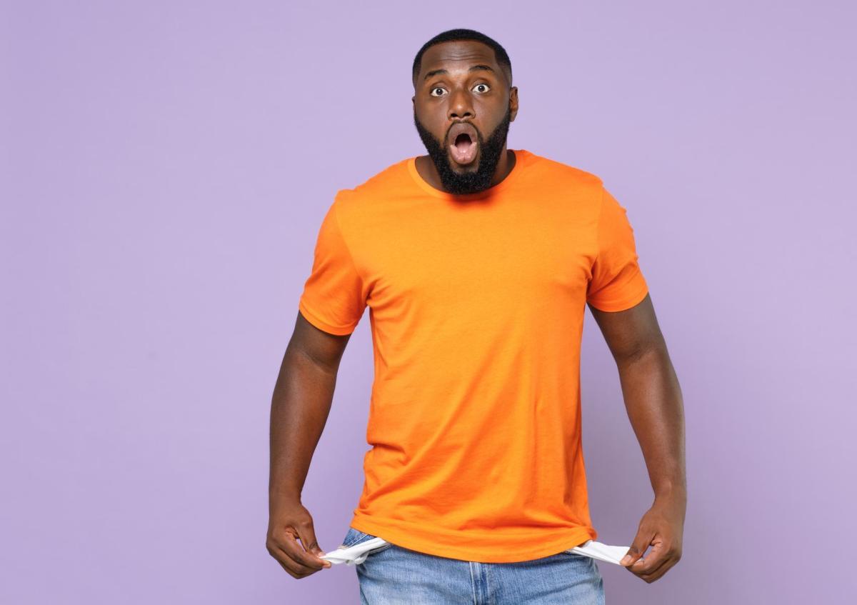 A surprised young man pulls out the empty pockets of his jeans, showing they contain no money. His mouth is open wide and his eyes are wide, expressing shock or disbelief