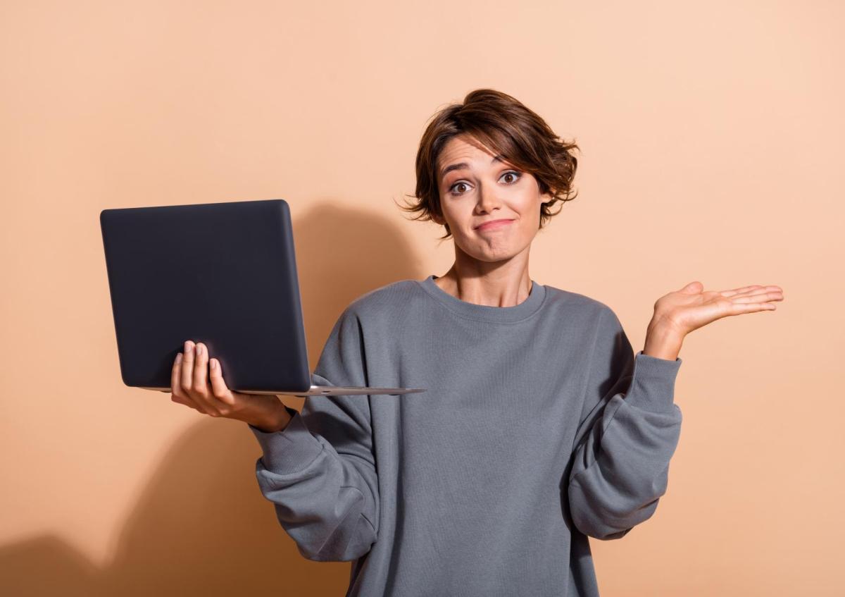 A young woman holding a laptop in one hand and raising her other hand with a shrug, wearing a confused or uncertain expression