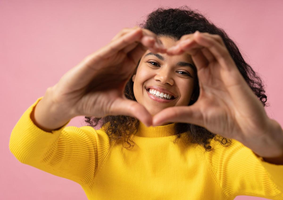 A cheerful young woman with curly hair, smiles warmly as she forms a heart shape with her hands in front of her face