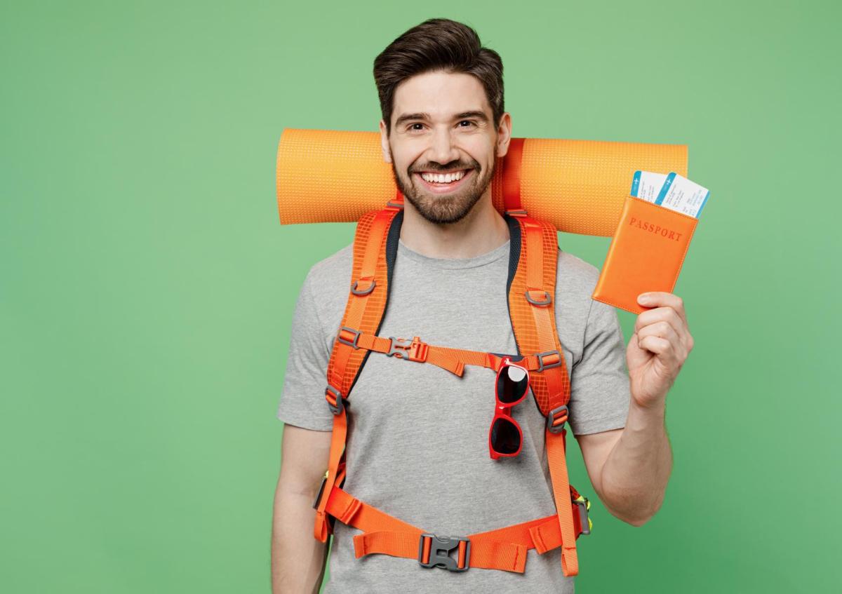 A young man with a beard, wearing a hiking backpack with a rolled-up sleeping mat, smiles broadly while holding a passport holder with boarding passes