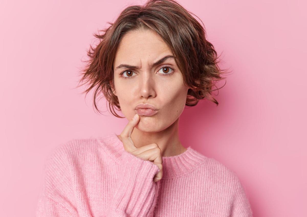 A young woman with an expression of confusion or puzzlement, with her eyebrows furrowed and her lips pursed. She is touching her chin lightly with her right index finger. 
