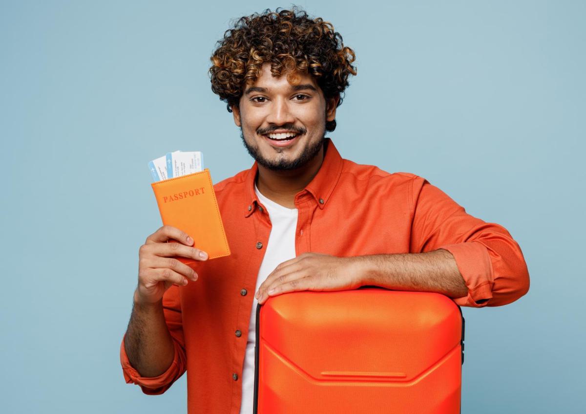 Man met gekruld haar en baard, glimlachend en een reiskoffer vasthoudend met zijn linkerarm. In zijn rechterhand houdt hij een paspoorthouder met vliegtickets erin.