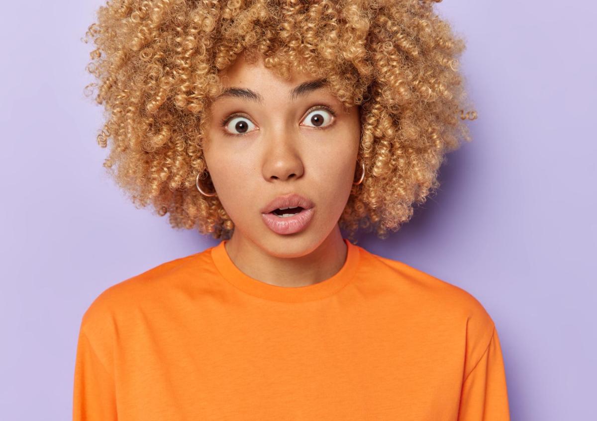 A young woman with curly hair wearing a bright top, looking directly at the camera with a surprised or shocked expression.