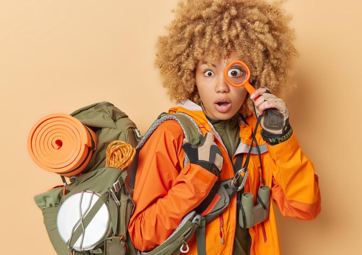 ChatGPT zei: A surprised woman dressed for outdoor adventure, carrying a backpack with camping gear and holding a magnifying glass up to one eye.