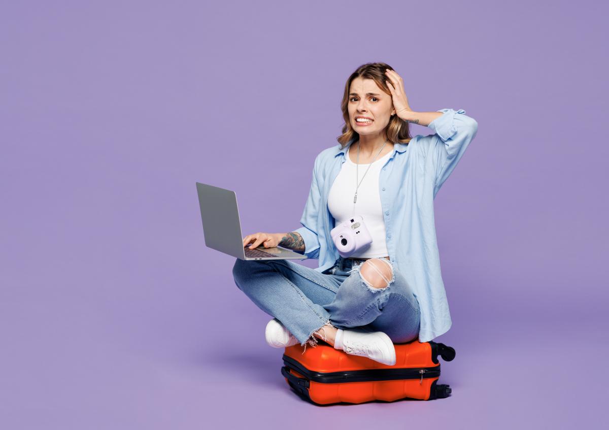 Vrouw zit op een koffer in een gekruiste houding met een laptop op haar rechterknie, haar linkerhand aan haar hoofd, en kijkt bezorgd of nerveus.