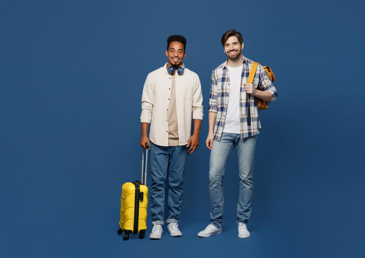 Two young men stand smiling, one holding a wheeled suitcase and the other carrying a backpack over one shoulder. Both appear ready for travel or an adventure