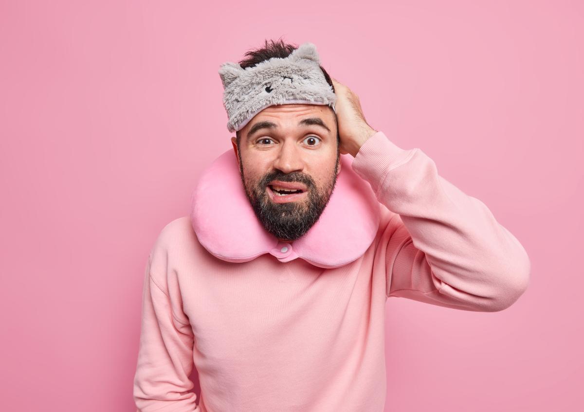A man looks confused or worried while wearing a travel neck pillow and a sleep mask pushed up on his forehead. He scratches his head with one hand
