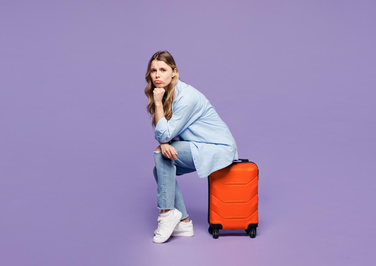 A young woman sitting on a suitcase, resting her chin on her hand with a thoughtful or slightly worried expression.