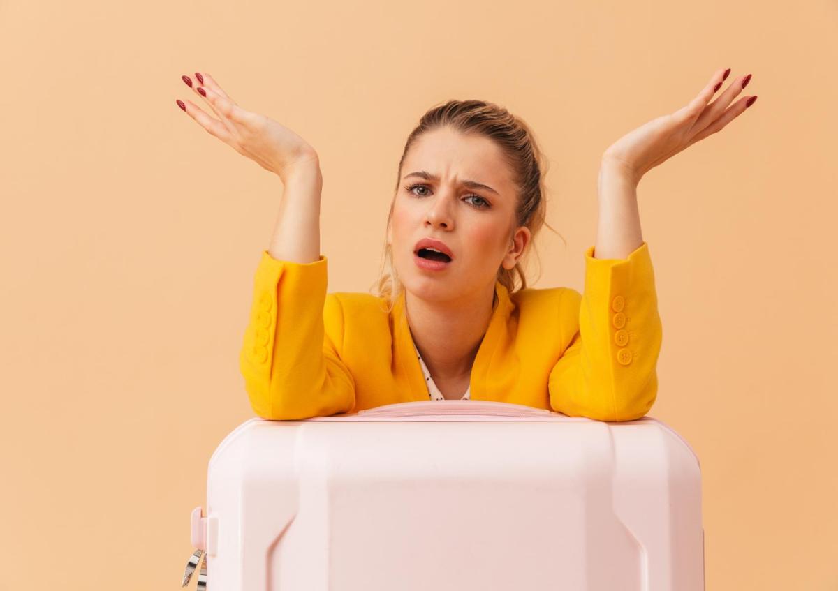 A frustrated woman leaning on a suitcase with her arms raised and mouth open, expressing confusion or annoyance.