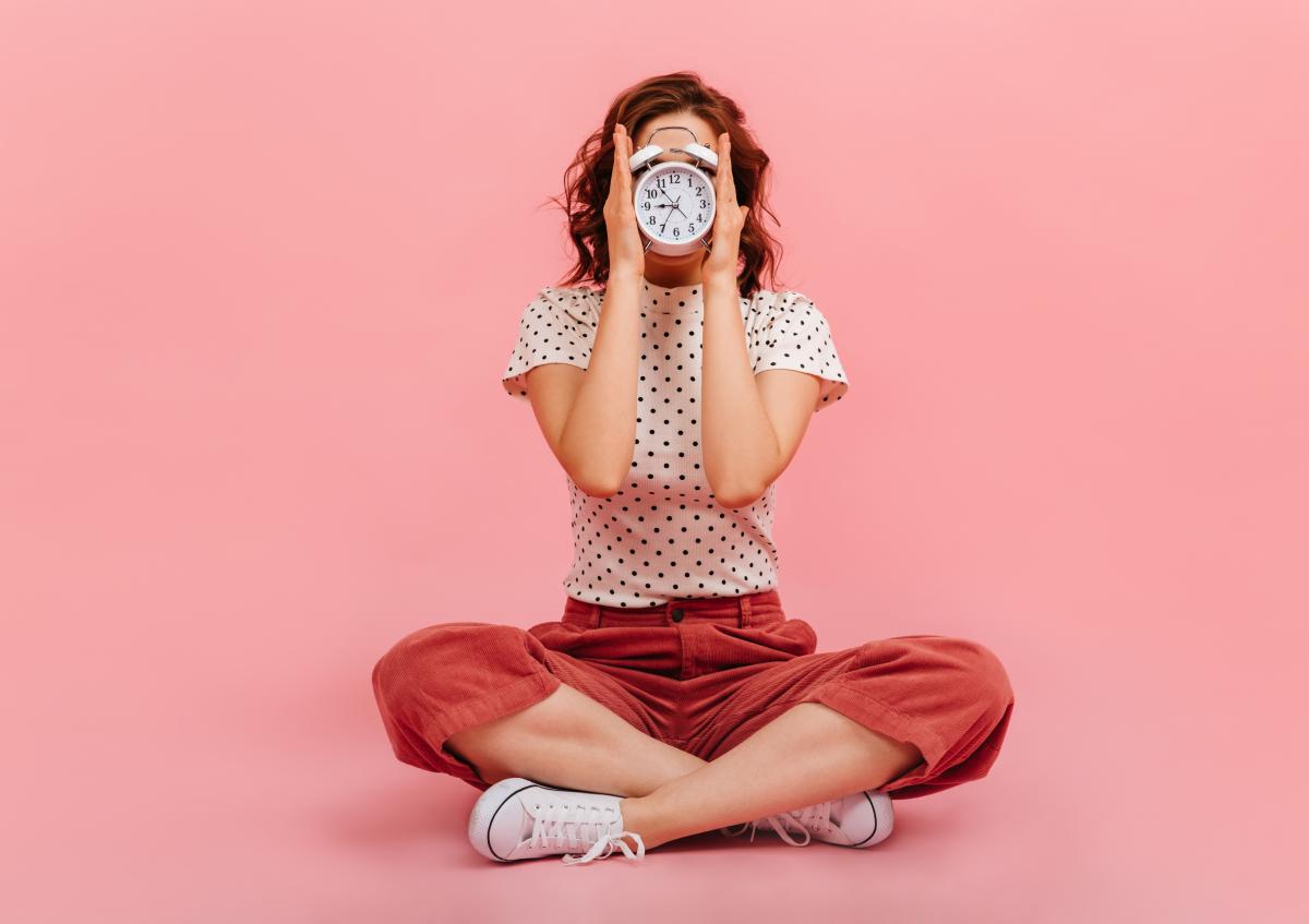 A young woman sitting cross-legged on the floor, holding a clock in front of her face, covering it completely.