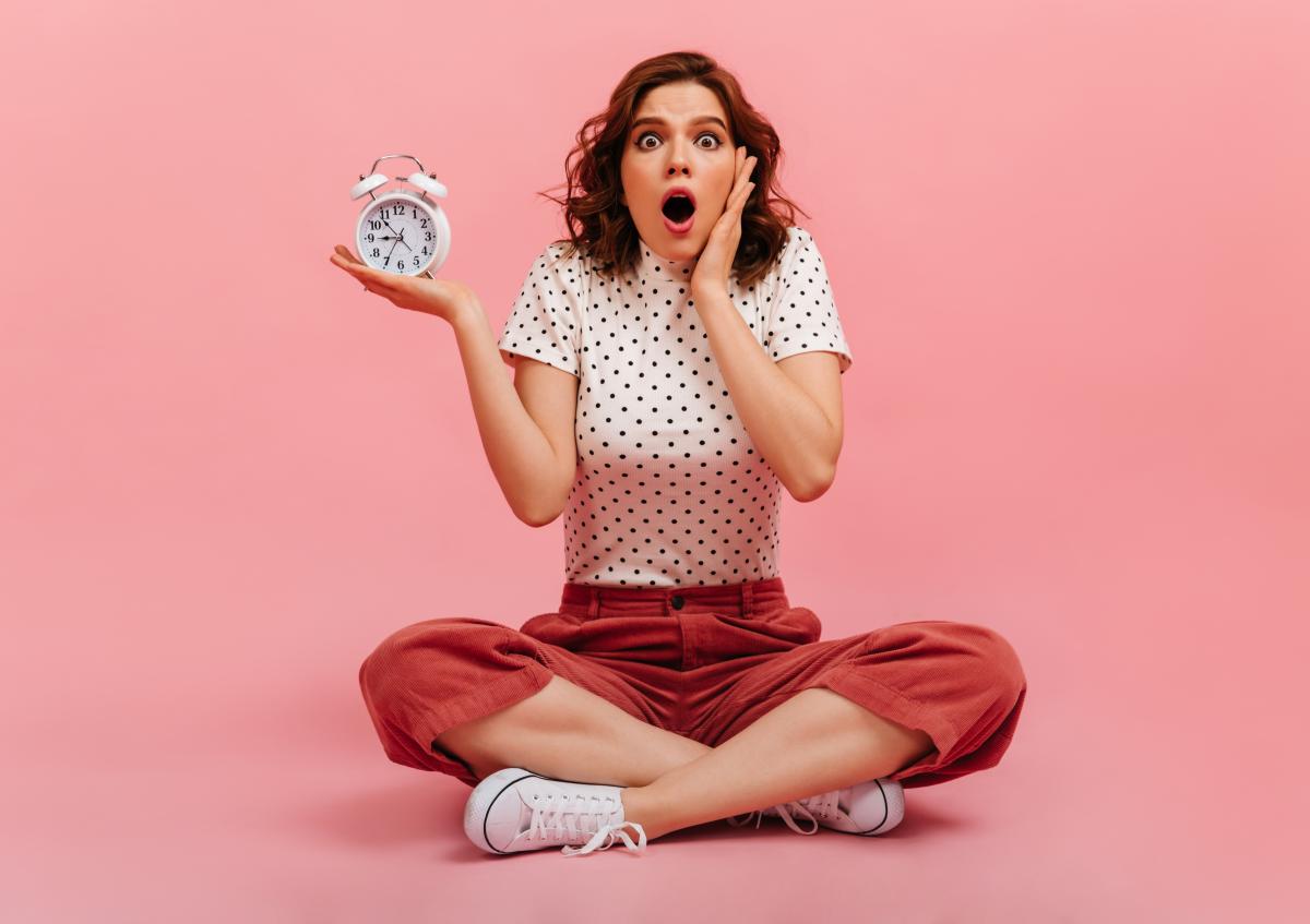 A young woman sitting cross-legged looks surprised while holding an alarm clock in one hand and supporting her head with the other.