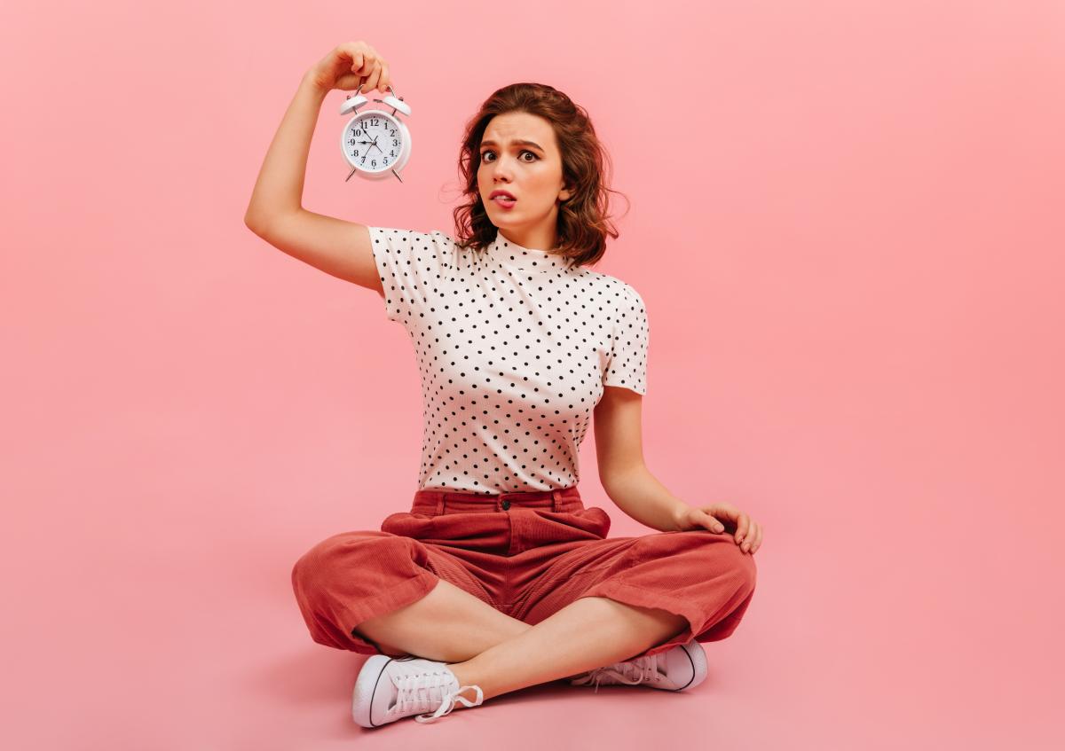 A young woman sitting cross-legged on the floor, holding an alarm clock up in one hand with a confused expression.