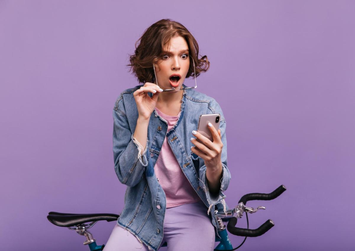 A young woman sitting on a bicycle, holding a smartphone in one hand, while removing her glasses with the other hand, showing a shocked or surprised expression.