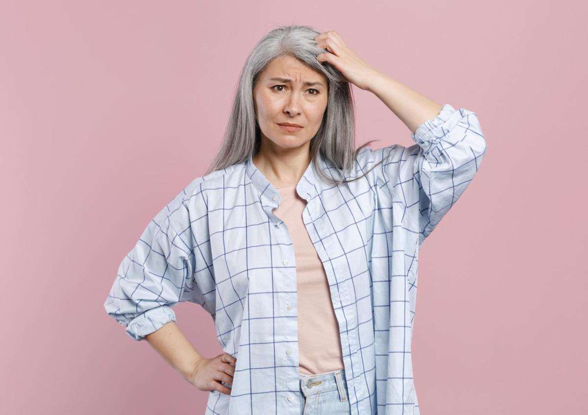 Een vrouw van middelbare leeftijd met lang haar staat met één hand op haar heup en krabt met de andere aan haar hoofd, met een verwarde of vragende blik.