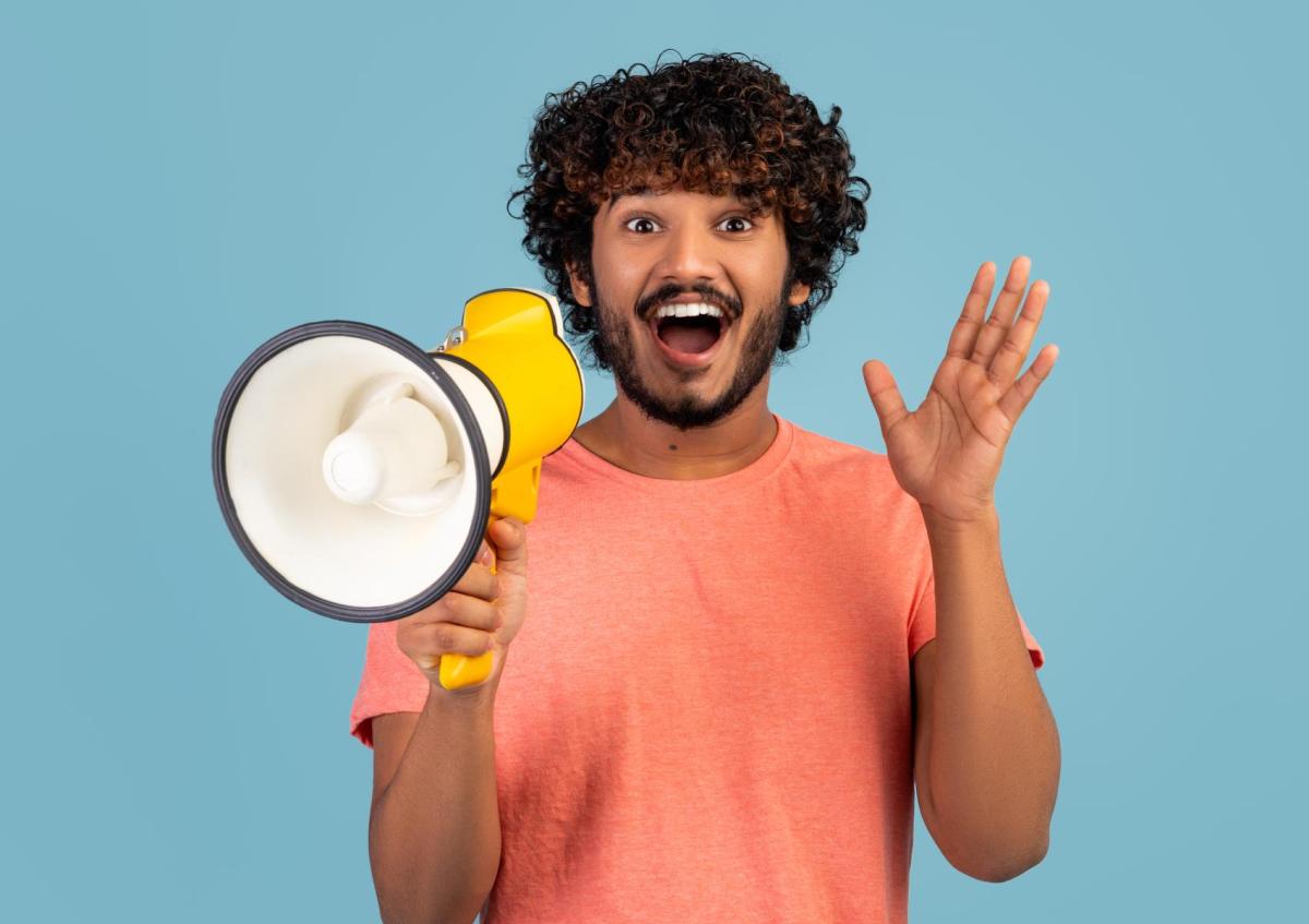 A young man with a curly hair and beard is holding a megaphone with one hand and waving with the other hand, looking excited and happy.