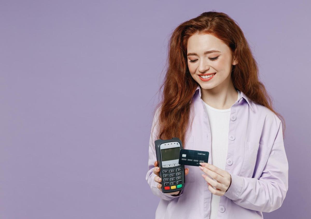 Een vrouw glimlacht terwijl ze in de ene hand een betaalterminal vasthoudt en in de andere een creditcard, klaar om een betaling te doen.