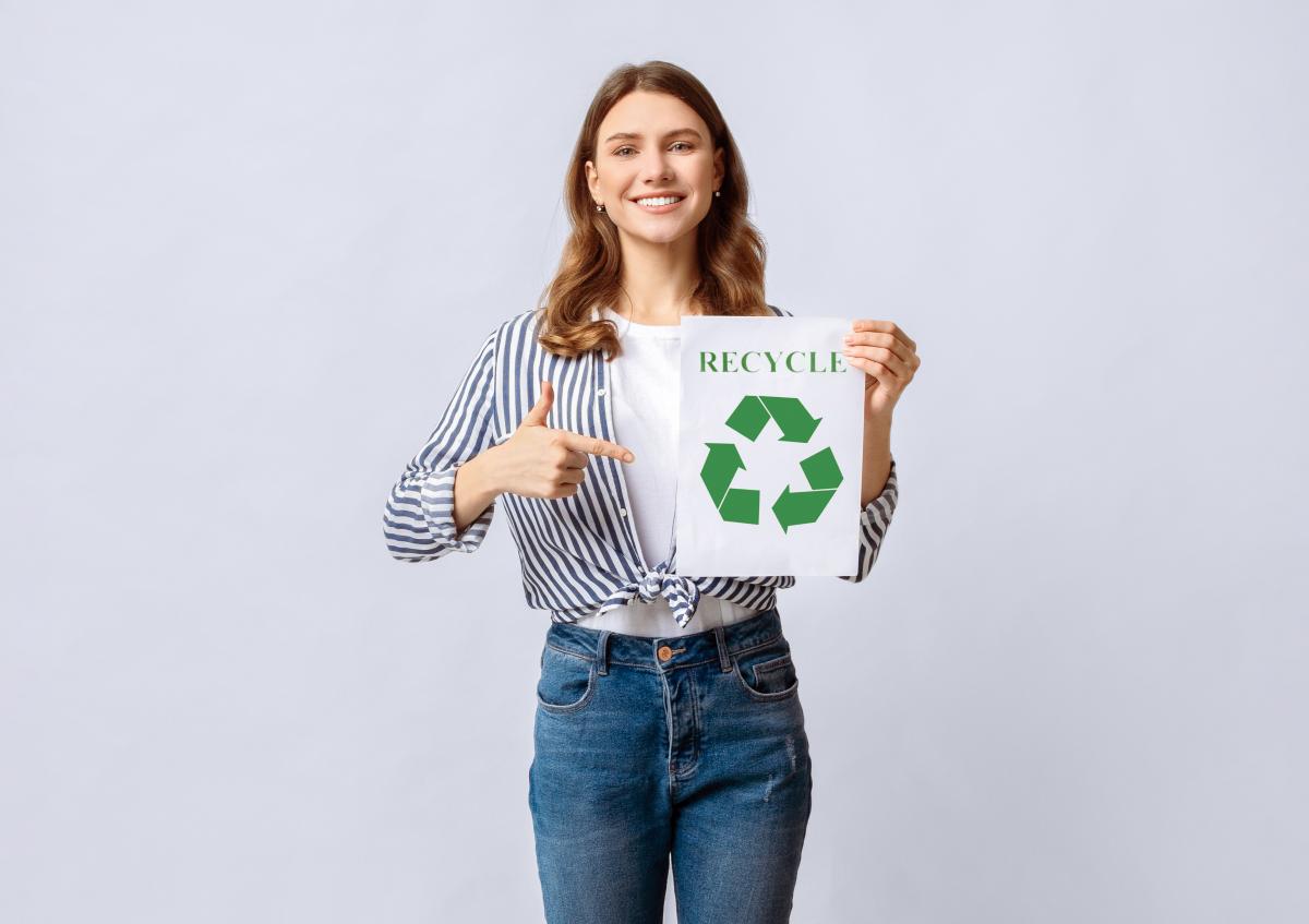 A smiling woman is holding a paper with a recycling symbol and pointing at it, expressing enthusiasm and encouragement for recycling.