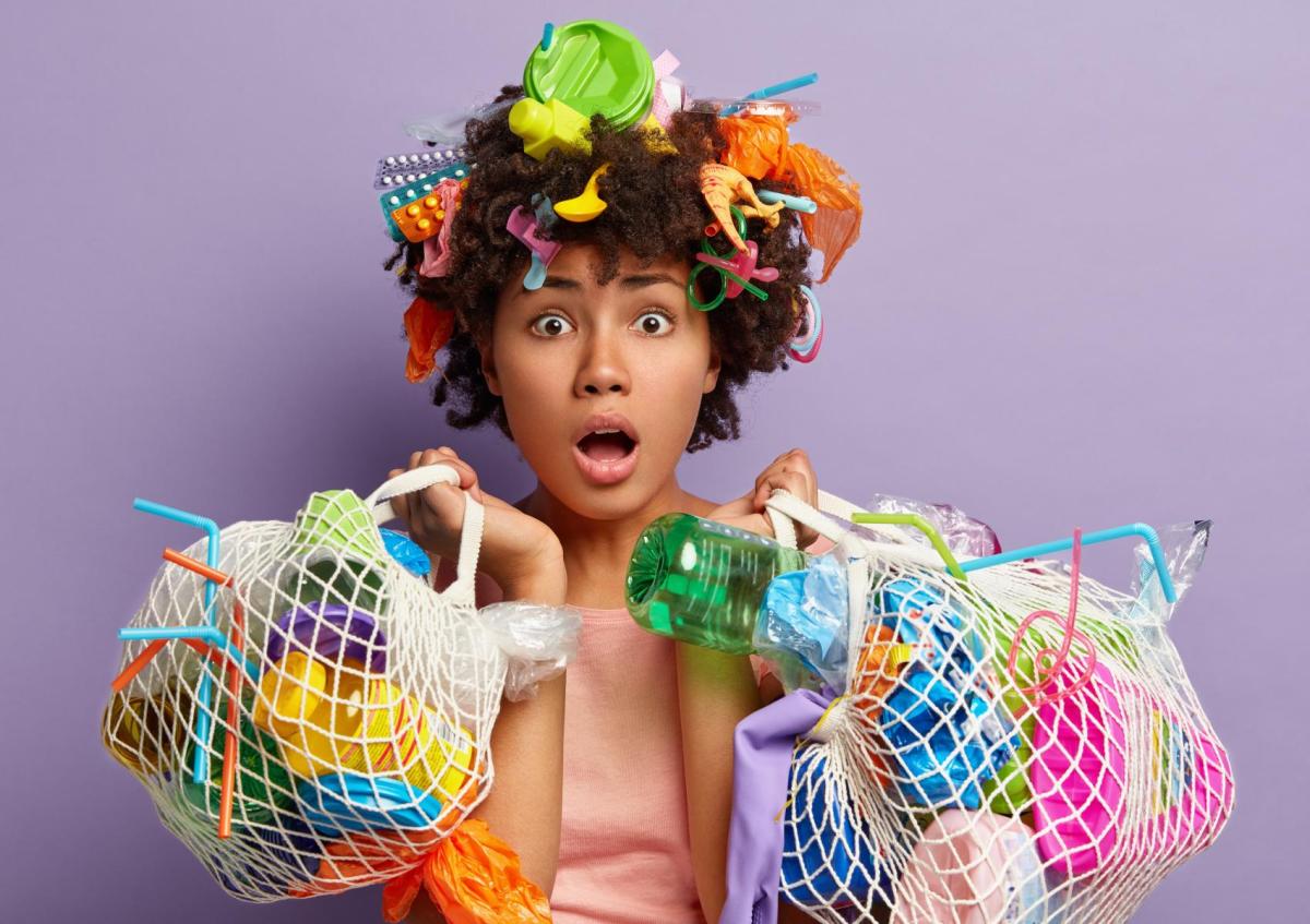 A surprised woman holding two mesh bags filled with various plastic items, while her hair is adorned with multiple small plastic objects, expressing shock or concern.