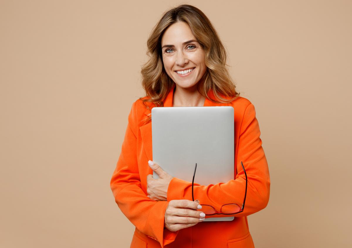 A woman holding a laptop close to her chest with one arm, while holding a pair of glasses in her other hand, smiling warmly at the camera.
