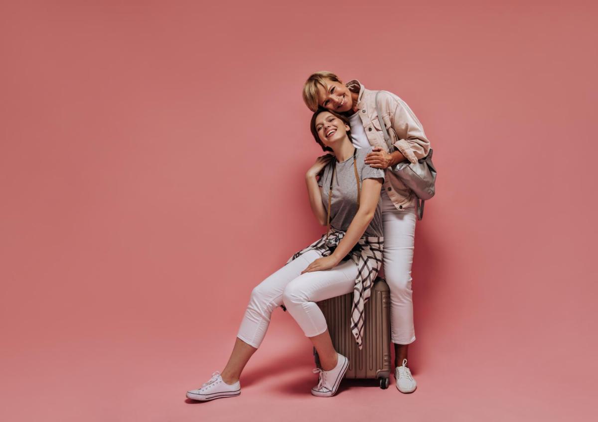 Twee vrouwen lachen blij en ontspannen. Eén zit op een koffer, de ander staat achter haar en leunt met haar hoofd tegen dat van de zittende vrouw. Ze omhelzen elkaar liefdevol.