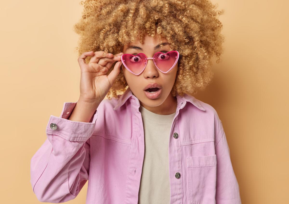 A young woman with curly hair and heart-shaped sunglasses looks surprised, holding one side of her glasses up as if to get a clearer view.