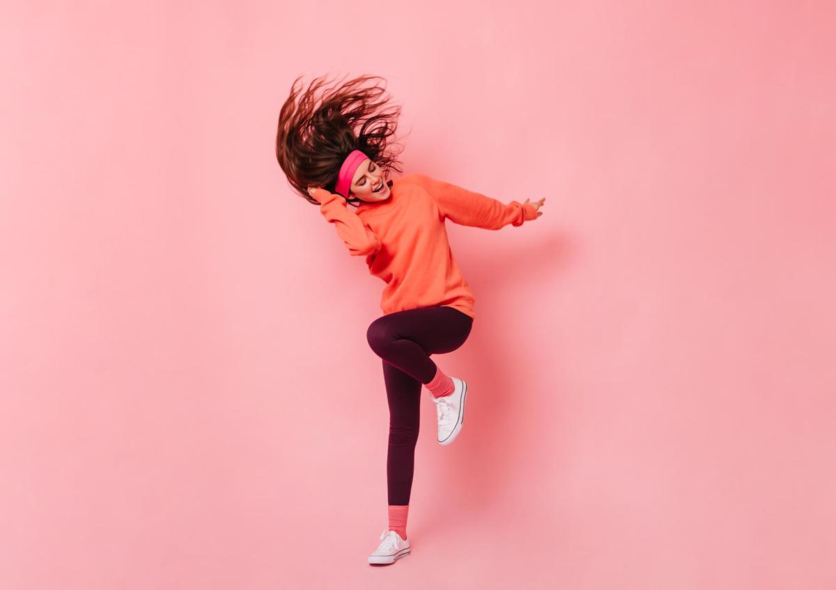A young woman energetically dances with one leg raised, hair flipping, eyes closed, and a joyful expression on her face.